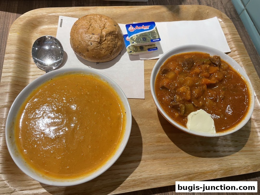 A wooden tray holds two bowls of soup, a bread roll, butter, and a spoon.