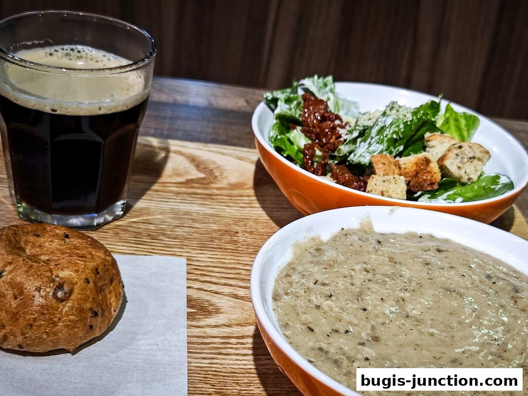 A glass of dark beverage, a bun, a salad, and a bowl of soup on a wooden table.