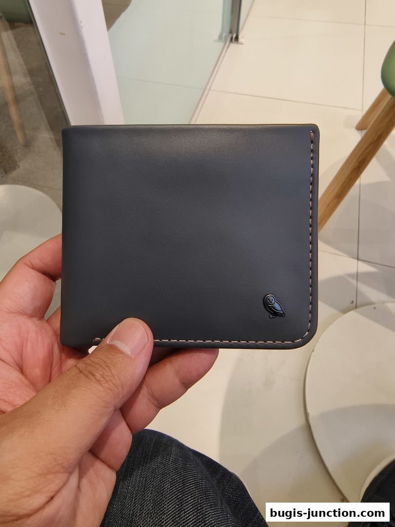 A hand holds a dark grey leather wallet with a small owl logo.