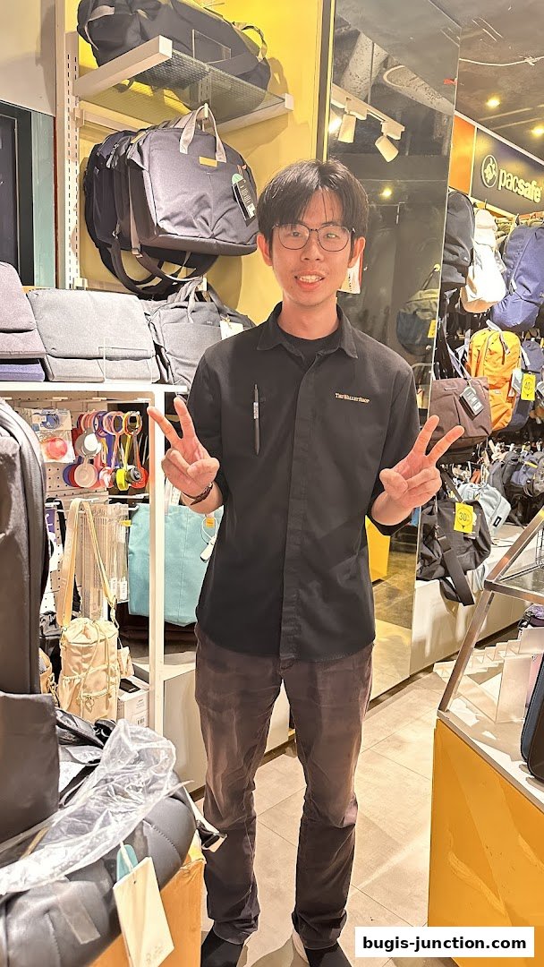 A smiling employee at The Wallet Shop poses with peace signs amidst a display of bags.