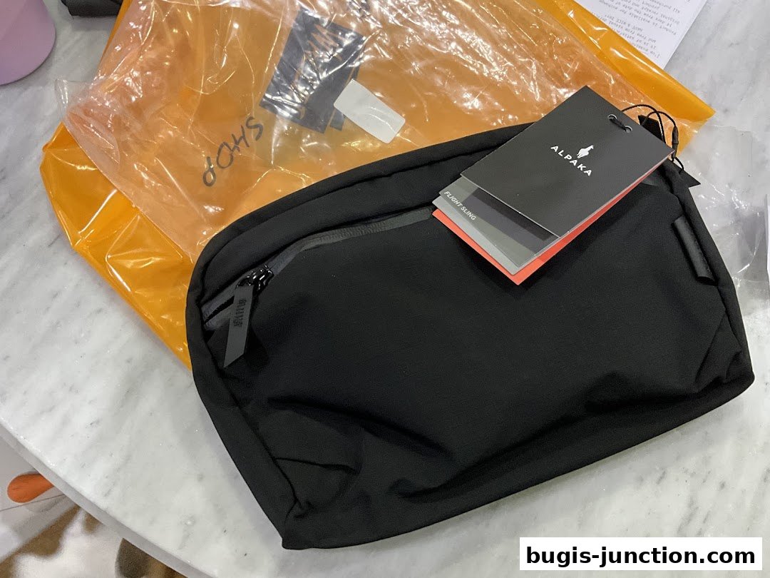 A black Alpaka Flight Sling bag with tags, placed on a marble surface next to an orange package.