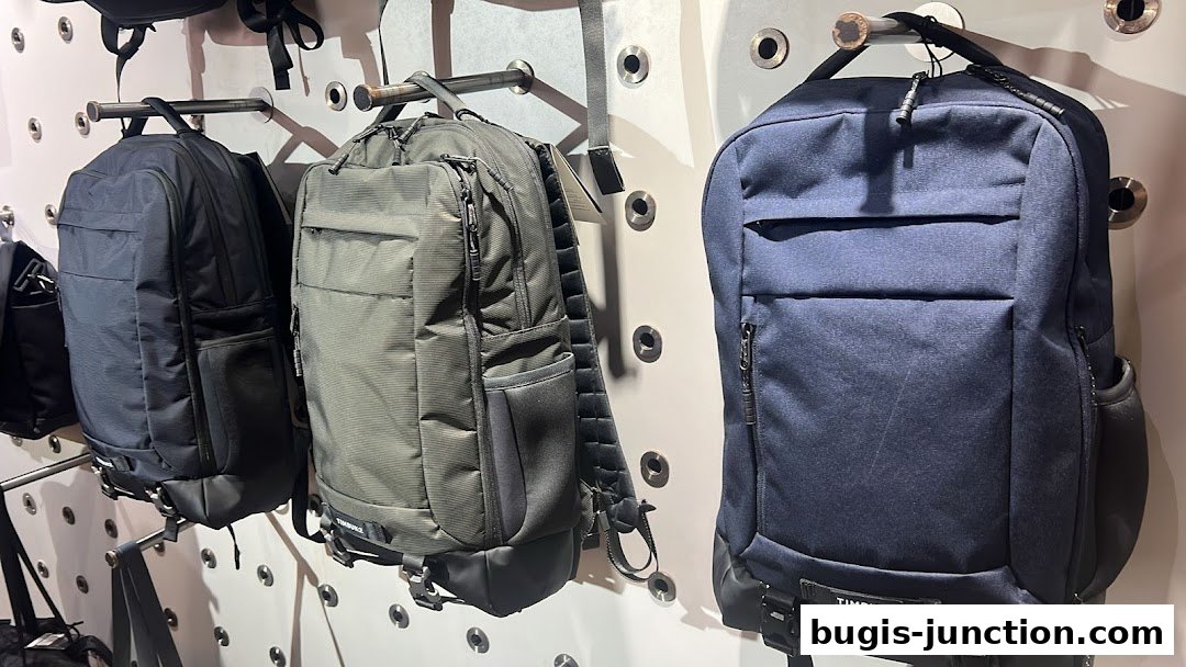 Three Timbuk2 backpacks displayed on a wall with metal hooks.