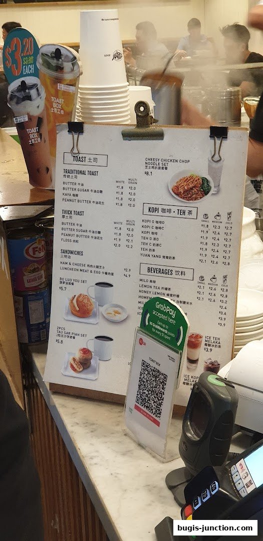 A menu board at Toast Box Bugis Junction displays various toast, sandwiches, kopi, teh, and beverage options.
