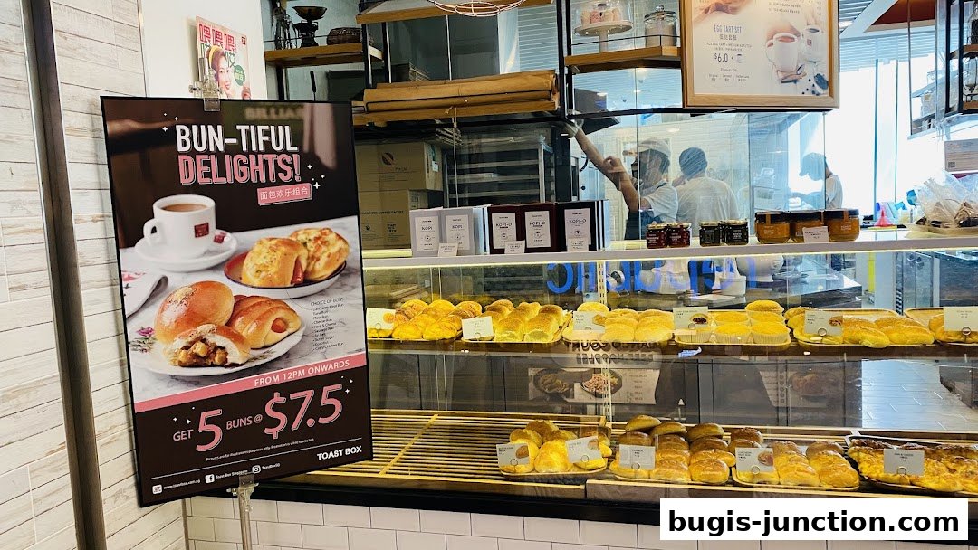 A promotional poster for Toast Box featuring a coffee and bun set, with a display of baked goods behind.