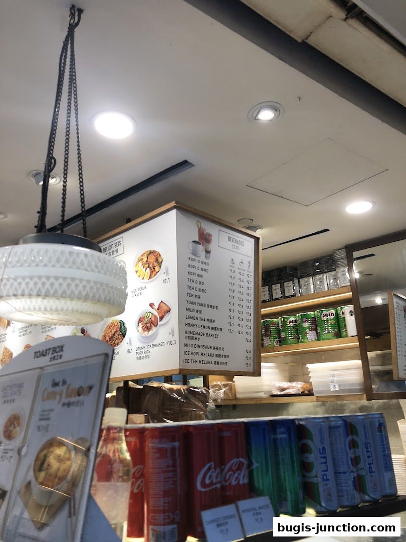 A menu board displaying beverages and meal sets at Toast Box, with canned drinks in the foreground.