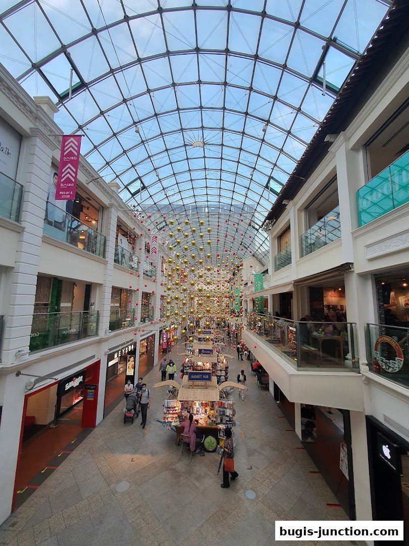 A wide shot of a shopping mall interior with a glass ceiling and various shops and stalls.
