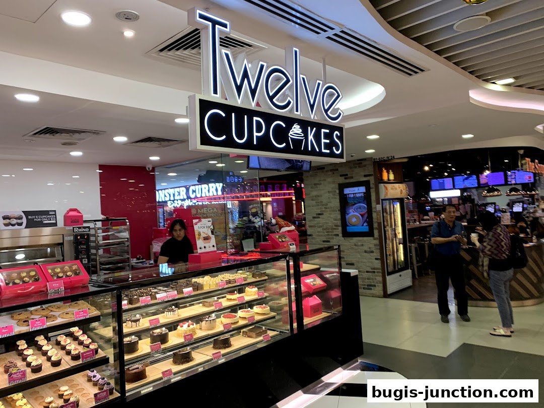 A brightly lit cupcake shop with a display of various cupcakes and cakes, and customers browsing.