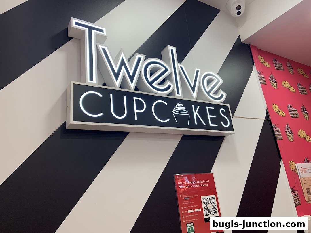 A brightly lit sign for 'Twelve Cupcakes' with a black and white striped wall and cupcake-themed wallpaper.