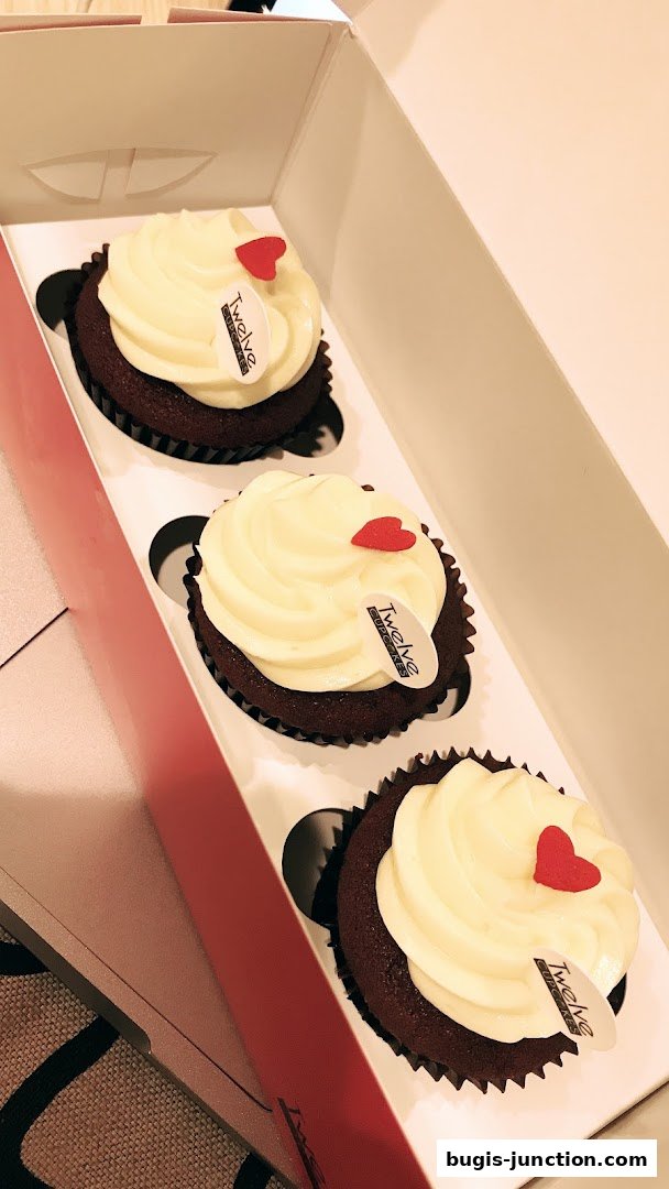 A box of three red velvet cupcakes with white frosting and red heart decorations.