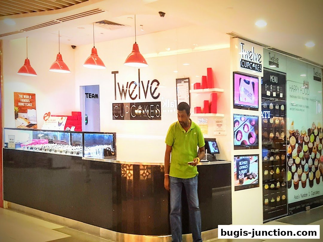 A man stands at the counter of a brightly lit cupcake shop with a display of cupcakes.