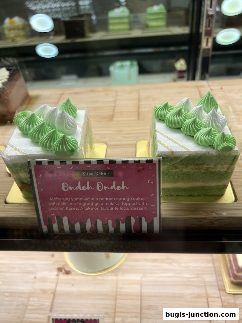 Two slices of Ondeh Ondeh cake with green and white frosting in a display case.