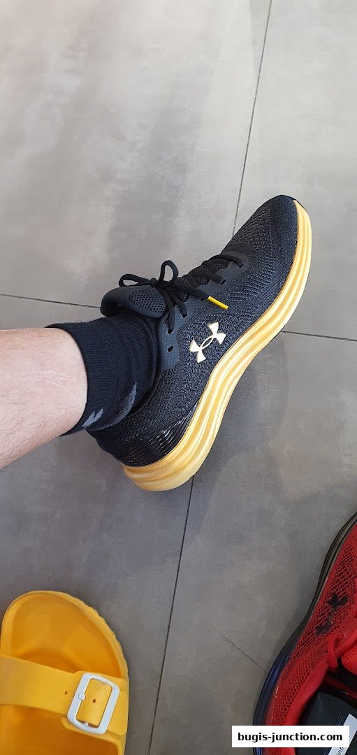 A person wearing black Under Armour sneakers with a yellow sole and yellow sandals.