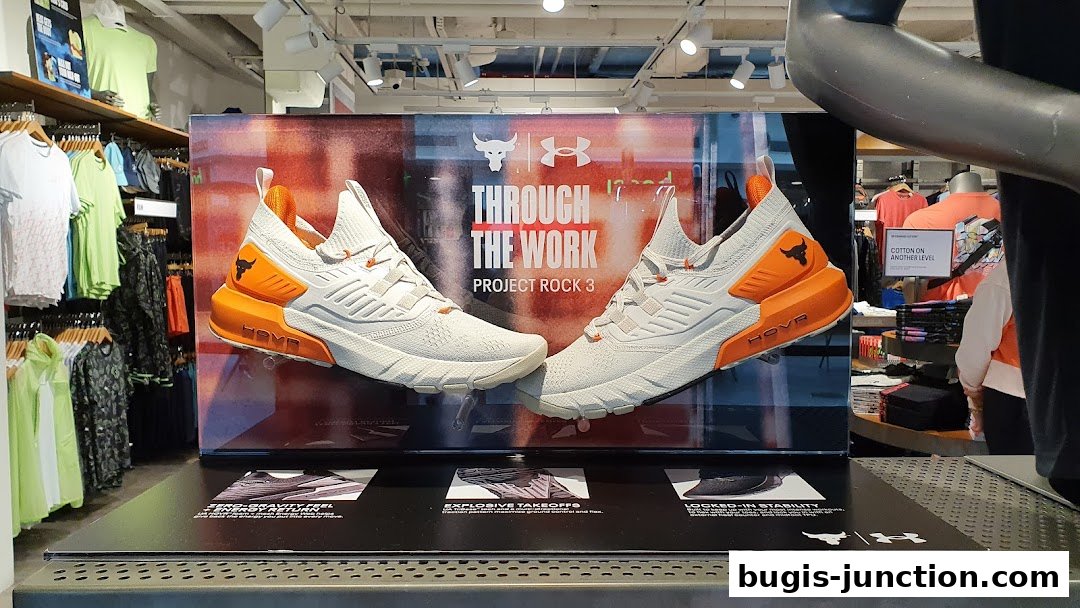 Under Armour Project Rock 3 shoes displayed with marketing graphics and apparel in the background.