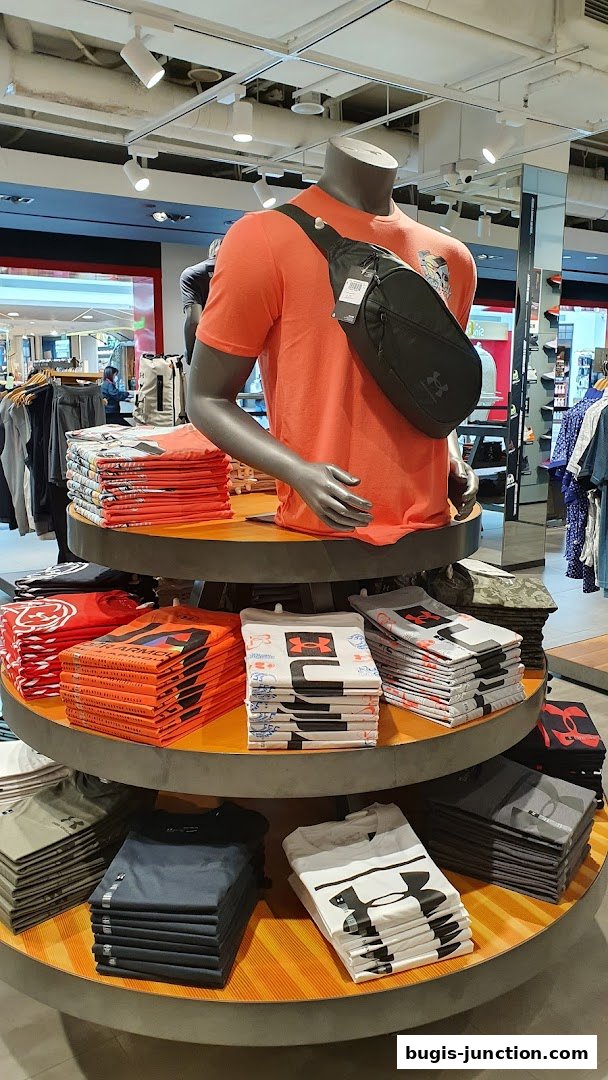 Mannequin wearing an orange t-shirt and a sling bag, surrounded by stacks of t-shirts.