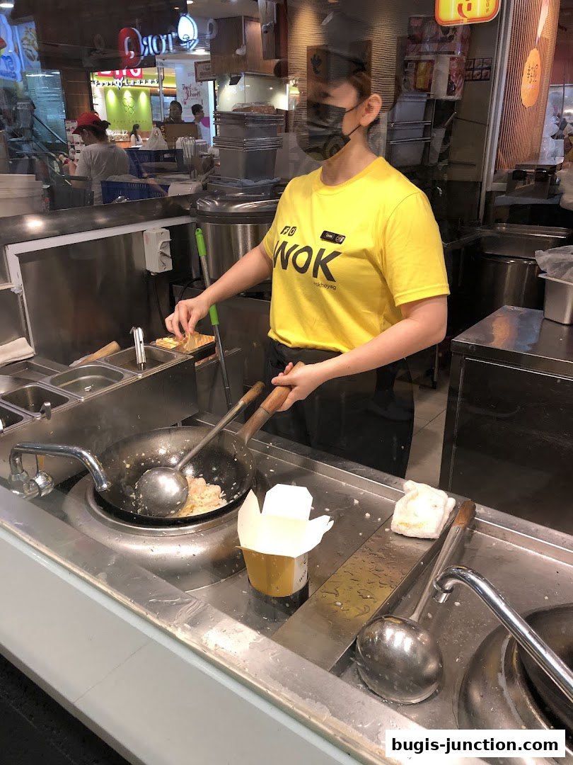 A chef in a yellow WOK t-shirt cooks food in a wok at a food stall.