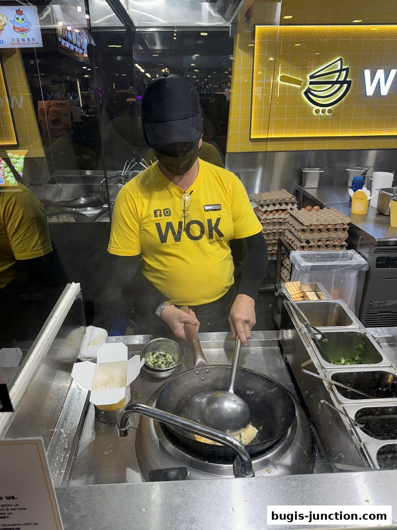 A chef in a yellow WOK shirt cooks food in a wok at a stall.
