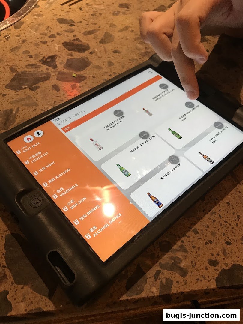 A hand is shown using a tablet to browse the menu of Xiao Long Kan Hotpot, specifically the alcohol drinks section.