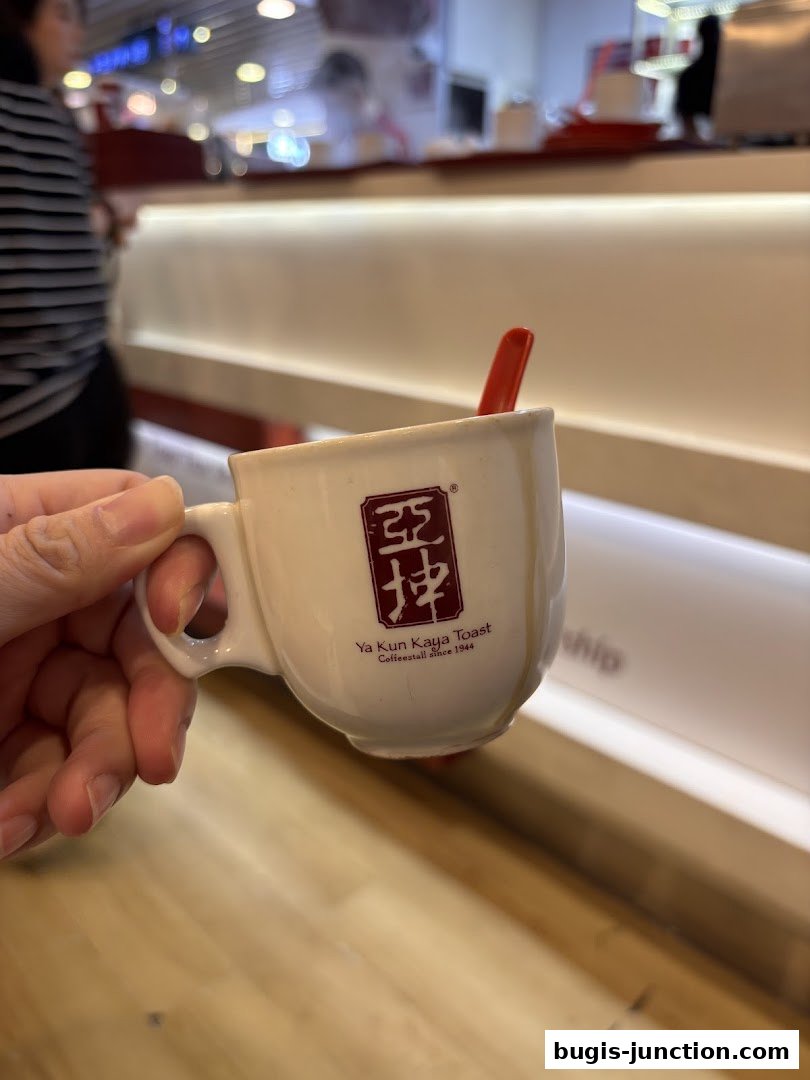 A hand holds a small white cup with the Ya Kun Kaya Toast logo and a red spoon.