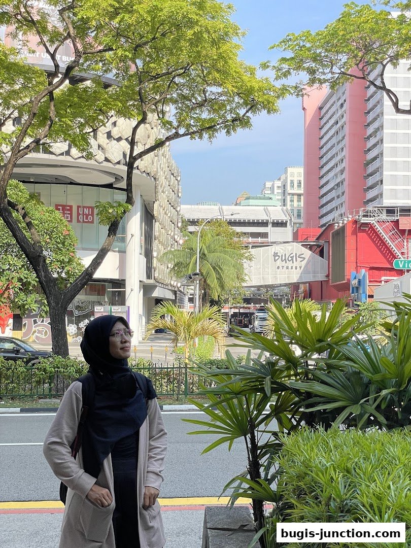 A woman stands on a sidewalk with lush greenery and buildings in the background.